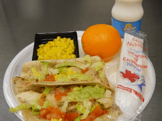 Chicken tacos in a whole grain tortilla with low-fat cheese, lettuce and tomato served with an orange, corn, and fat-free milk